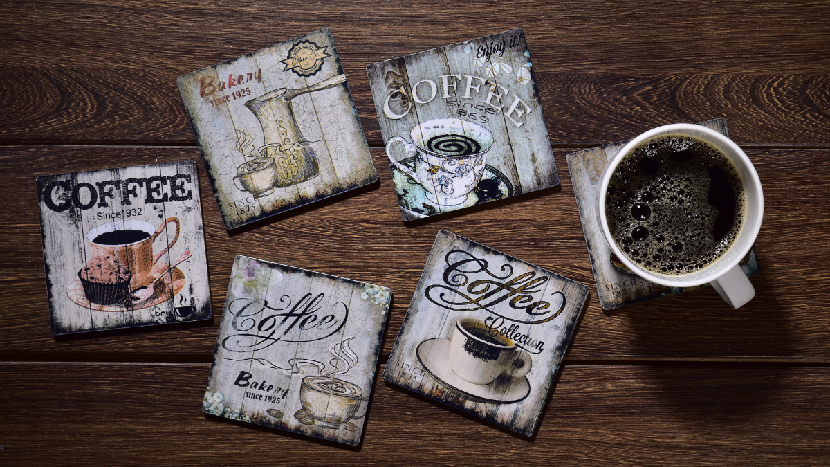 A set of vintage-style square coffee-themed coasters with rustic designs scattered on a dark wooden surface, alongside a cup of black coffee placed on one of the coasters.