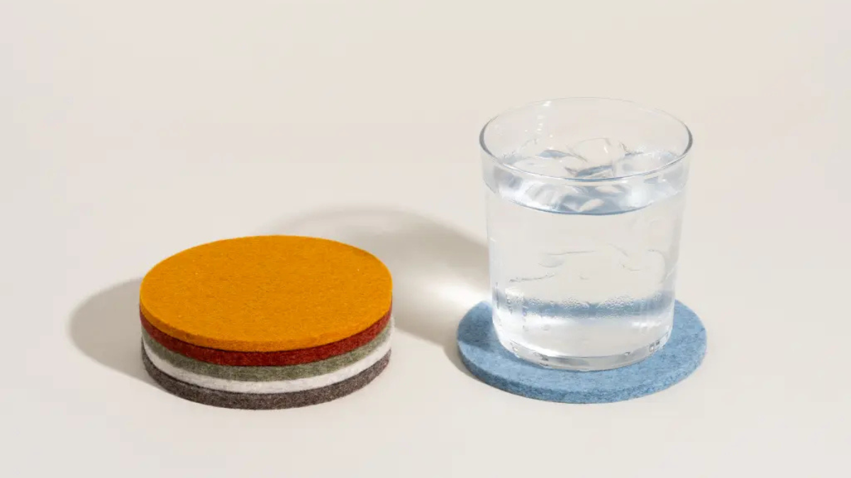 A set of round felt coasters in mustard, brown, white, and gray stacked neatly, with a glass of water placed on a blue coaster beside them.