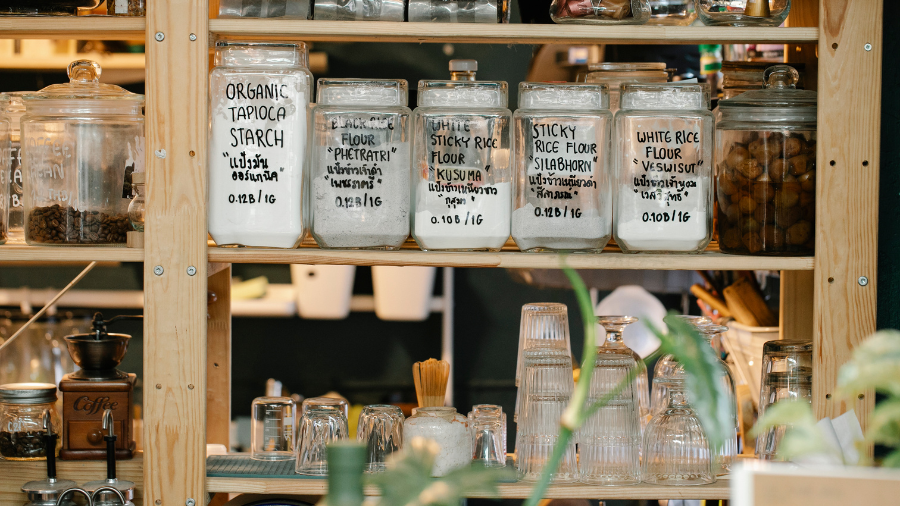 Wooden kitchen shelf with glass jars labeled with different types of flour and starch, along with neatly arranged glassware and kitchen items.