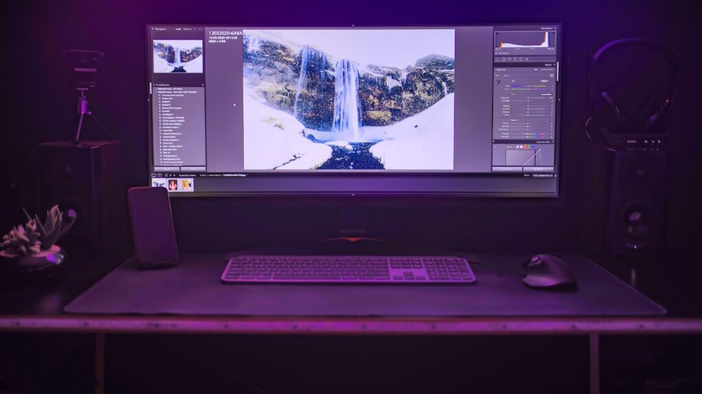 A modern editing setup with a widescreen monitor displaying photo editing software, a keyboard, a mouse, and a dark-themed workspace with purple lighting.